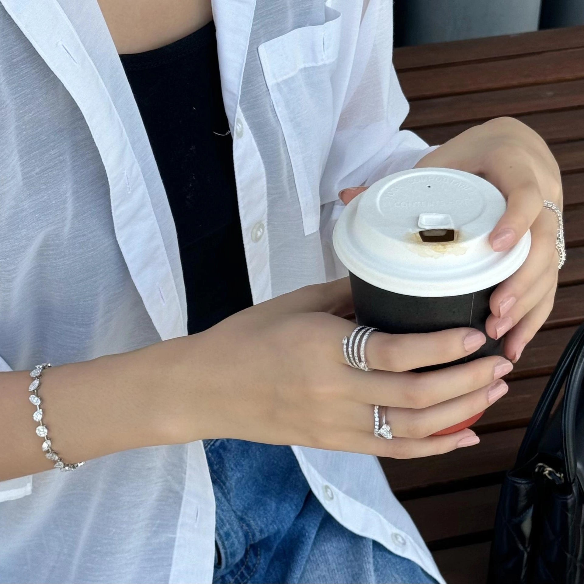 Wearing multiple diamond rings holding a coffee cup outdoors on a wooden bench.