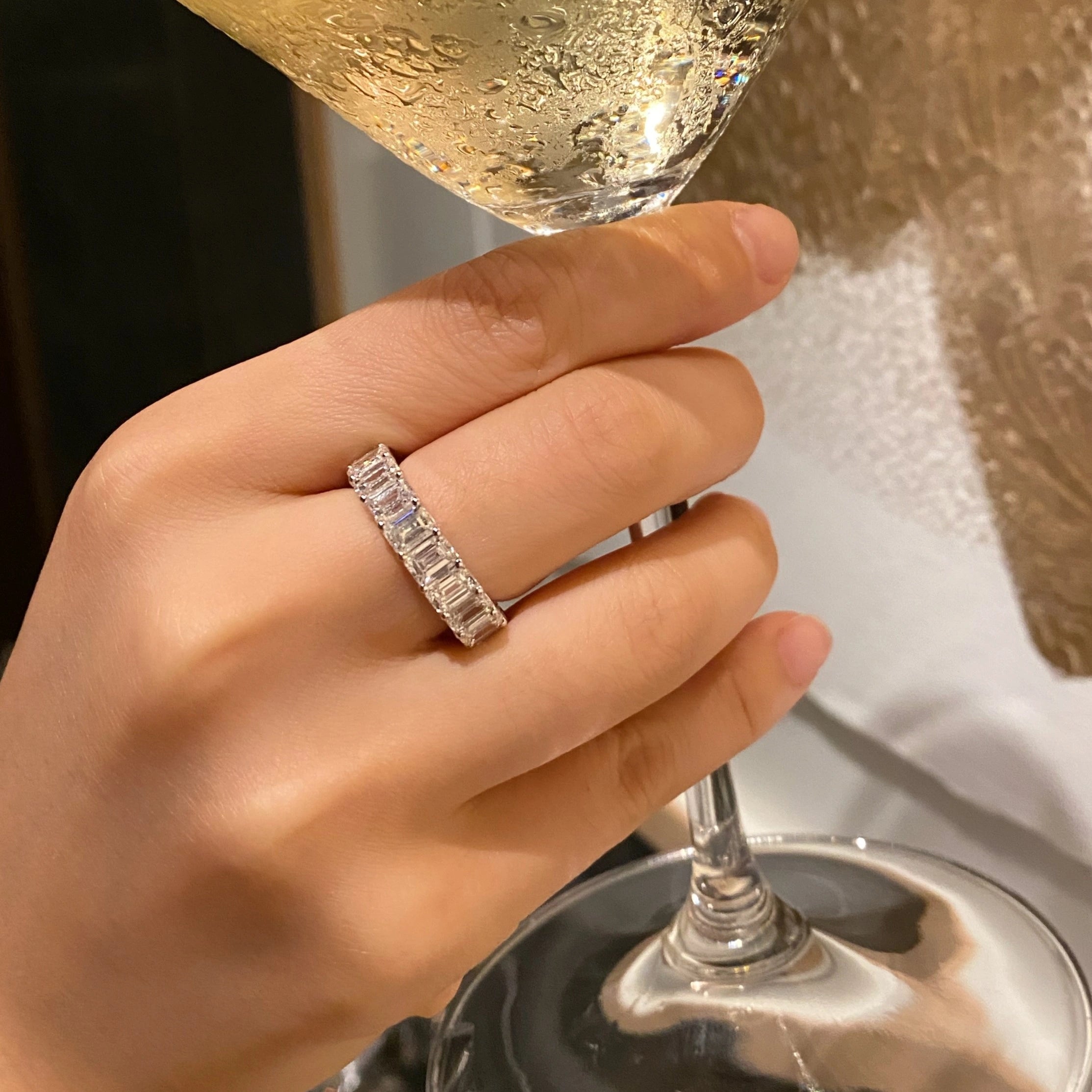 Hand wearing an emerald cut eternity ring holding a glass of champagne